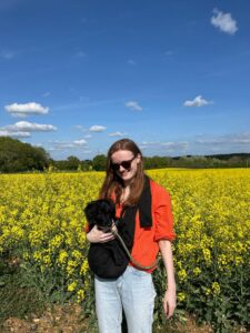 Picture of Lydia (Regular Giving Officer) stood in front of a field of yellow flowers and blue sky carrying her dog, lemon, in a bag.