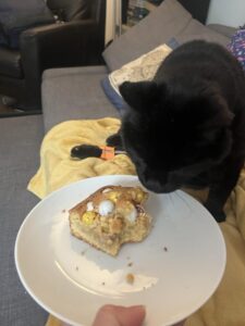A picture of a piece of partially eaten cake on a plate with a black cat leaning over to investigate!
