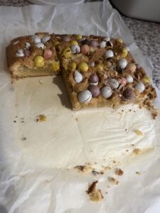 A picture of a half eaten cake in baking paper with mini eggs on top of the cake.
