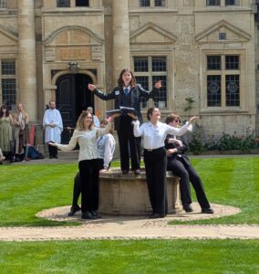 A picture of students performing in the Medieval Mystery Cycle. A female student stands on top of the well at St Edmund Hall with stood on the floor below, all with their arms aloft.