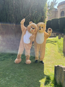 Two teddy bear mascots stood in St Edmund Hall graveyard in the sunshine cheering with their arms in the air