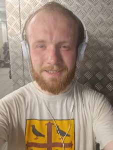 A selfie of Timothy Powell wearing headphones with a metal background in a Teddy Hall Freshers T-Shirt taken after cycling for the 1317 Challenge